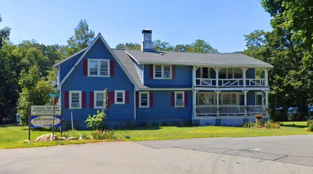 A two-story blue house with white trim and red shutters, featuring a covered porch on both the first and second floors. The house is surrounded by green grass and trees, with a sign in front that reads 'Lakeside Care Home Inc'.