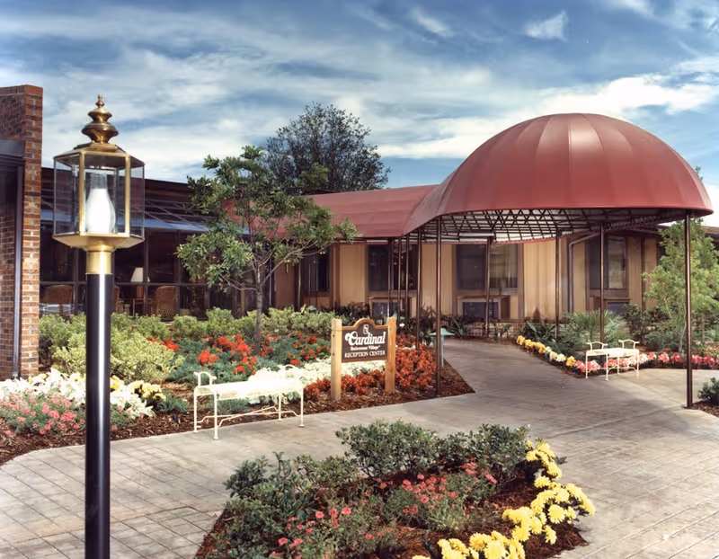 Entrance to Cardinal Village Reception Center with a red canopy over the walkway, surrounded by landscaped gardens with colorful flowers and greenery. There are white benches along the path and a vintage-style lamp post in the foreground.