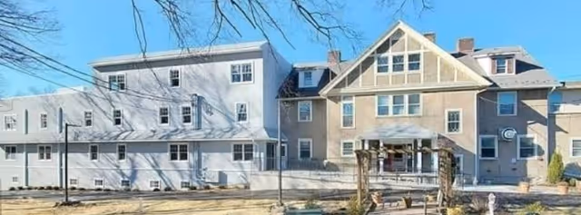 Exterior view of a multi-story senior living facility building with beige and white walls, multiple windows, and a small covered entrance area. The foreground shows a paved area and some landscaping with bare trees under a clear blue sky.