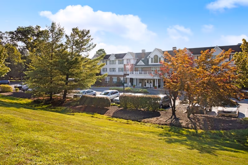 Exterior view of Brighton Gardens of West Orange, a multi-story senior living facility with a brick and white facade, surrounded by trees with green and autumn-colored leaves, a parking lot with several cars, and a grassy lawn in the foreground under a partly cloudy blue sky.