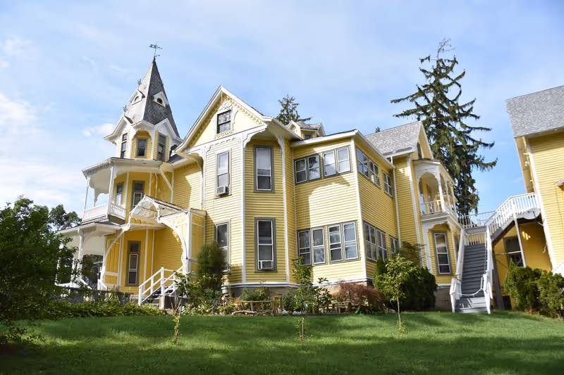 Yellow Victorian-style multi-story building with a turret, porches, and exterior staircases on a green lawn.