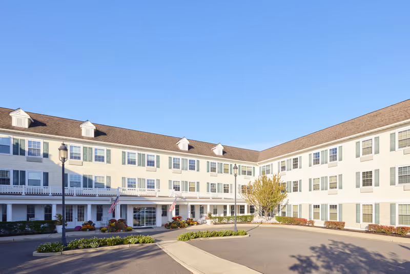 Exterior view of a three-story senior living facility building with white siding, green shutters, and a brown roof under a clear blue sky. The entrance is decorated with flowers and American flags, and there are two street lamps and landscaped greenery in front.