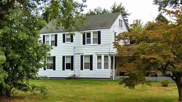 A two-story white house with black shutters surrounded by green trees and a grassy lawn under a clear sky.