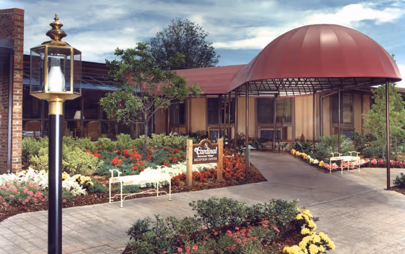 Front entrance of Cardinal Village reception center with a red dome canopy, landscaped flowerbeds, benches, and a lamp post.