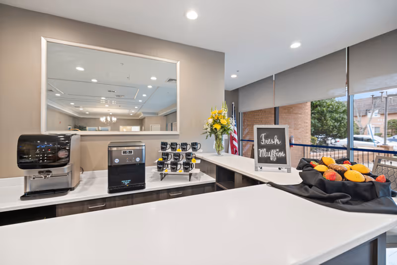 A bright and clean coffee and snack station with a coffee machine, a hot water dispenser, a rack of black mugs, a vase of yellow flowers, and a basket of fresh muffins on a white countertop. A small chalkboard sign reads 'Fresh Muffins'. Large windows with roller shades reveal an outdoor view with trees and buildings.