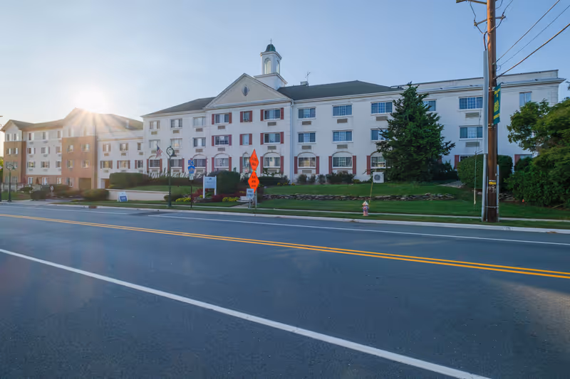 Exterior view of a large, multi-story senior living facility building with white brick walls and red window shutters. The building has a small cupola on the roof and is surrounded by well-maintained landscaping including bushes and flowers. The sun is low in the sky, creating a lens flare effect. There are road signs and a fire hydrant visible along the street in front of the building.
