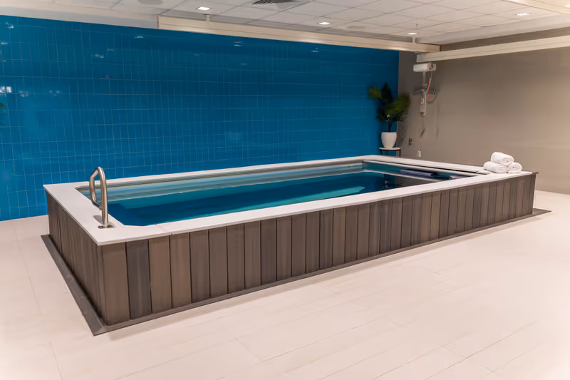 Indoor therapy pool with a wooden panel exterior, blue tiled wall in the background, a metal handrail on the left side, and a stack of white towels on the right corner. There is a potted plant on a small table near the back wall.