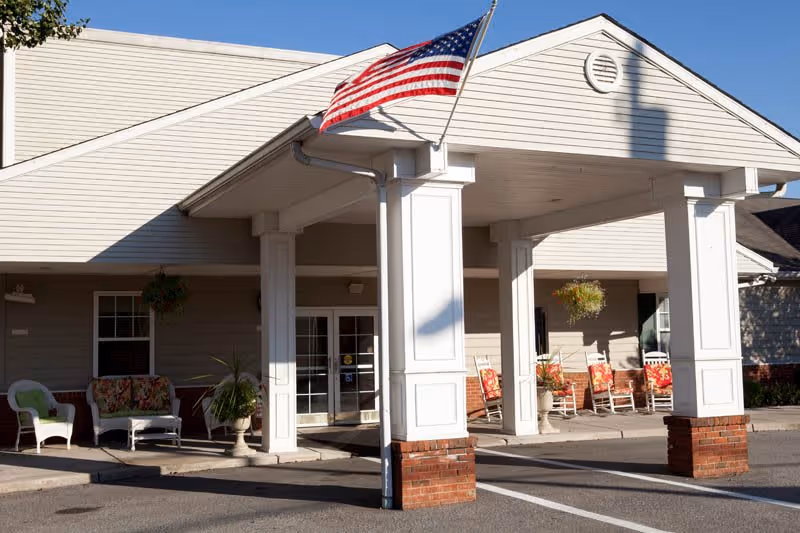 Entrance of a senior living facility with a covered drop-off area supported by white columns with brick bases. An American flag is mounted on one of the columns. There are several chairs and potted plants arranged near the entrance under the covered area.