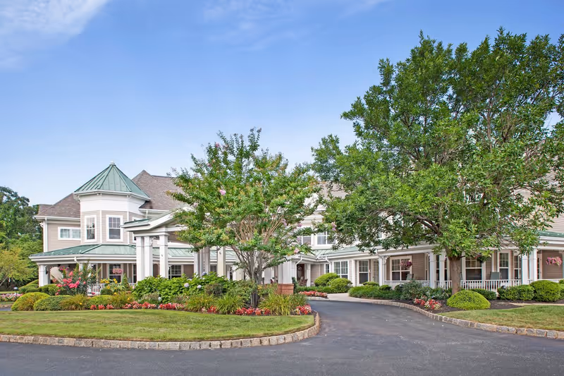 Exterior view of a senior living facility named Sunrise of Wall, featuring a large building with a green metal roof, white columns, and a well-maintained garden with trees, shrubs, and colorful flowers. The driveway curves around a landscaped island in front of the entrance.