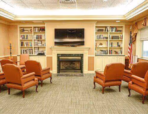 Cozy common room with armchairs arranged facing a fireplace and wall-mounted TV flanked by built-in bookshelves.