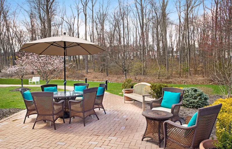 Outdoor patio area with a round glass table surrounded by six wicker chairs with turquoise cushions and a large beige umbrella overhead. Nearby, there is a wooden bench and additional wicker chairs with turquoise cushions. The patio is paved with bricks and is surrounded by green grass, flowering trees, and leafless trees in the background.