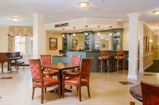 A bright and spacious common area with a small bar counter and stools, a wooden table with four red cushioned chairs, a piano in the corner, and decorative wall art. The area has light-colored walls and flooring, with ceiling lights and a sign above the bar that reads 'BON APPETIT'.