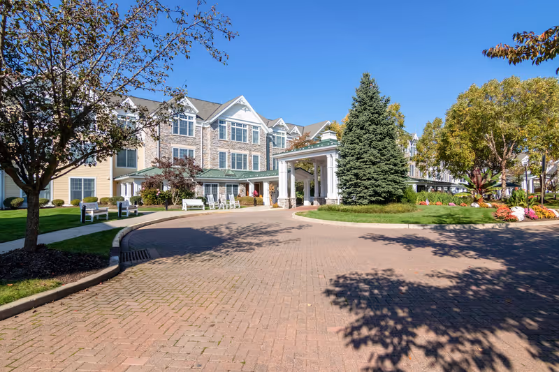 Exterior view of Sunrise of Cresskill senior living facility showing a large, multi-story building with stone and siding facade, a covered entrance with white columns, landscaped grounds with trees, shrubs, and flowers, and a brick-paved driveway under a clear blue sky.