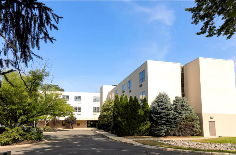 Front exterior of a three-story pale beige senior living building with landscaped trees and a driveway under a clear blue sky.