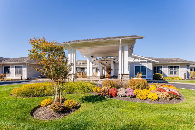 Exterior view of Sunrise of Jackson senior living facility with a covered entrance, manicured green lawn, colorful flower beds, and clear blue sky.