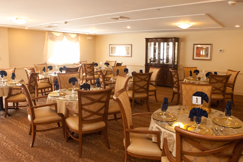 A dining room with multiple round tables covered with beige tablecloths, each set with gold chargers, silverware, glasses, and navy blue folded napkins. The room has beige walls, carpeted floors, a window with curtains, framed artwork, and a wooden china cabinet.