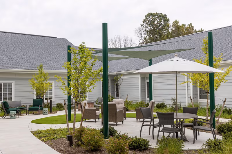Outdoor courtyard area with patio furniture including chairs and tables, some shaded by umbrellas and shade sails, surrounded by small trees and shrubs, with a building in the background.