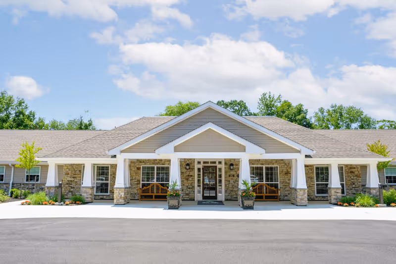 Front exterior view of a single-story senior living facility building with stone and beige siding, a gabled roof, two wooden benches on the porch, potted plants, and a clear blue sky with some clouds.