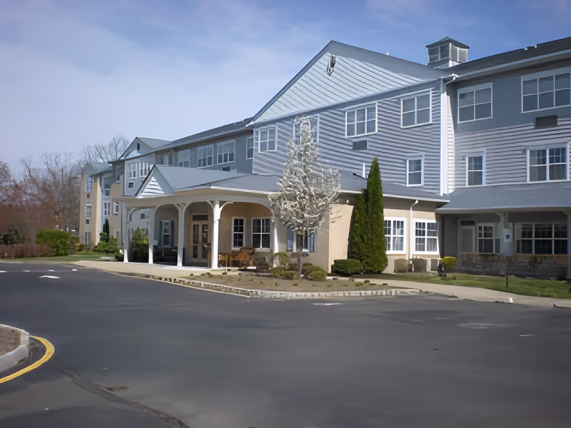 Exterior view of Magnolia Gardens, a multi-story senior living facility with white siding and multiple windows. The building features a covered entrance with white columns, surrounded by small landscaped bushes and trees. The sky is clear and blue.
