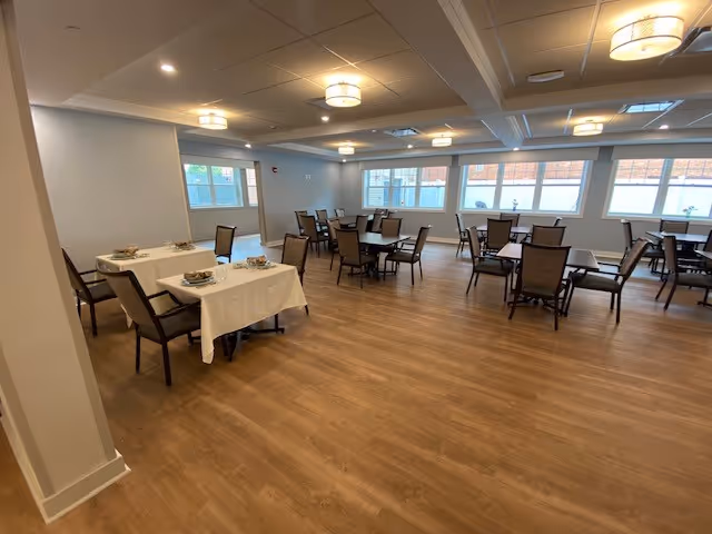 A bright communal dining room with multiple tables and chairs on wood-look flooring beneath recessed ceiling lights and windows.