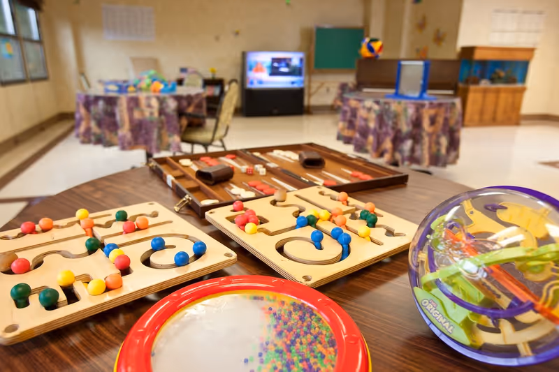 A recreational room with various tabletop games including wooden puzzles with colorful pegs, a backgammon set, and a transparent spherical maze puzzle on a wooden table. In the background, there are round tables covered with patterned tablecloths, chairs, a television, a chalkboard, and a fish tank.