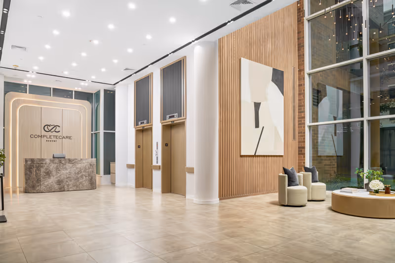A modern and spacious lobby area of Complete Care at Regent featuring a reception desk with the facility's logo on the wall behind it, two elevators with wooden paneling, a large abstract painting on a wooden slatted wall, and a seating area with two round chairs and a circular coffee table near large floor-to-ceiling windows.