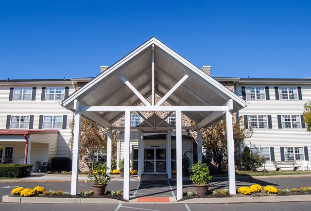 Front exterior view of Cherry Hill Senior Living facility with a large white covered entrance supported by pillars, potted plants, and yellow flowers along the driveway under a clear blue sky.