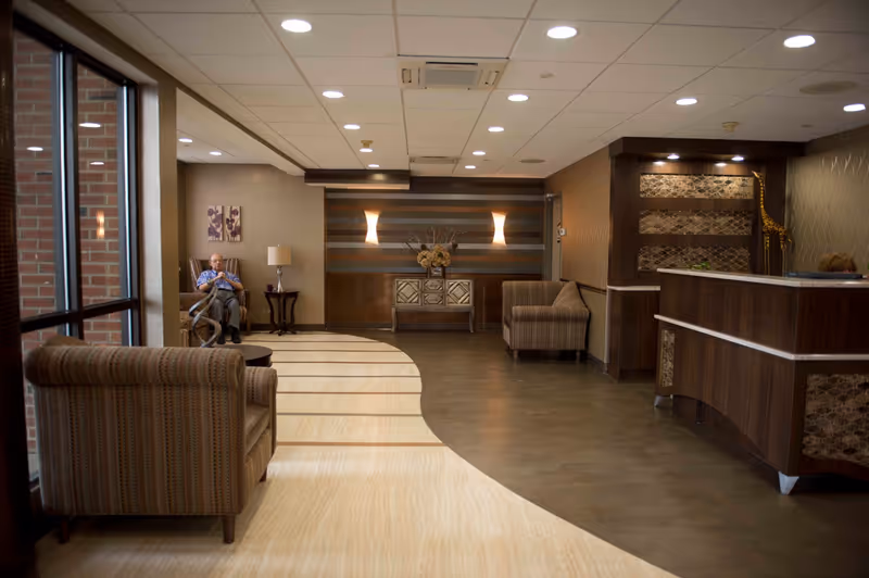 A spacious and well-lit reception area in Elmora Hills Healthcare & Rehabilitation Center featuring a wooden reception desk on the right, comfortable upholstered chairs, a small table, decorative wall lights, and a man sitting on a chair near the window on the left side.