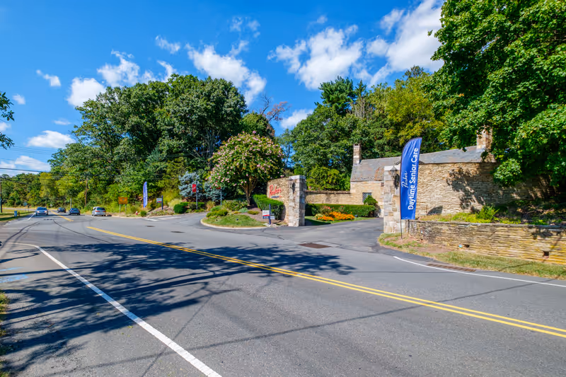 Road leading to the stone-gated entrance of Parker at Stonegate with landscaping and a blue 'Daytime Senior Care' banner.