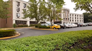 Exterior view of a multi-story healthcare and rehabilitation center building with a parking lot in front, several parked cars, and trees surrounding the area.