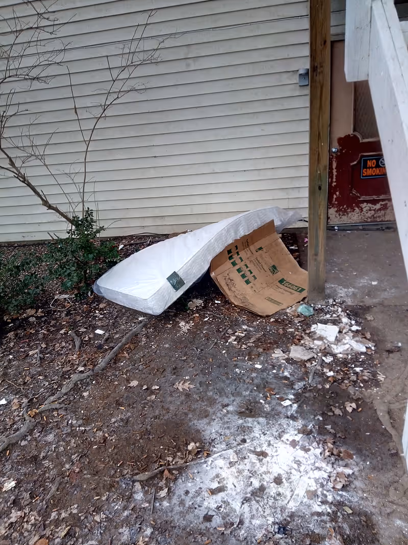 A discarded mattress and cardboard box lean against the exterior siding of a building beside a doorstep with scattered debris on the ground.