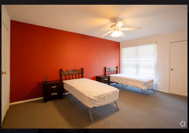 A bedroom with two single beds placed parallel to each other against a red accent wall. Each bed has a wooden headboard and is covered with a white sheet. Between the beds are two dark wooden nightstands with drawers. A ceiling fan with a light is mounted on the ceiling. A large window with closed blinds is on the right wall, and a closed white door is visible on the far right side of the room. The floor is carpeted in a neutral color.