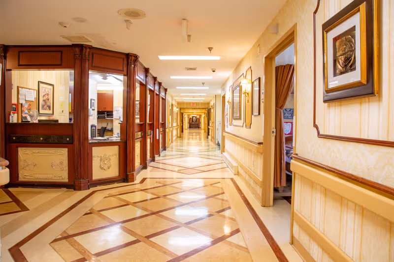 Ornately decorated, well-lit long hallway in a senior living facility with a reception desk on the left and framed artwork along the walls.