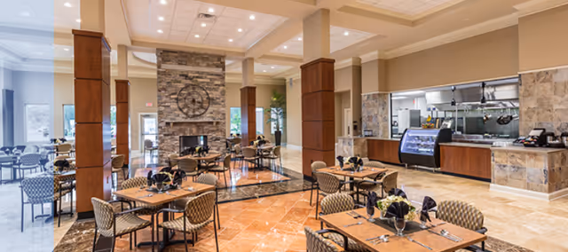 A spacious dining area in a senior living facility with multiple tables and chairs arranged neatly. The room features a stone fireplace with a large clock above it, wooden columns, and a serving counter with kitchen equipment visible behind it. The floor is tiled with a mix of light and dark tiles, and the ceiling has recessed lighting.