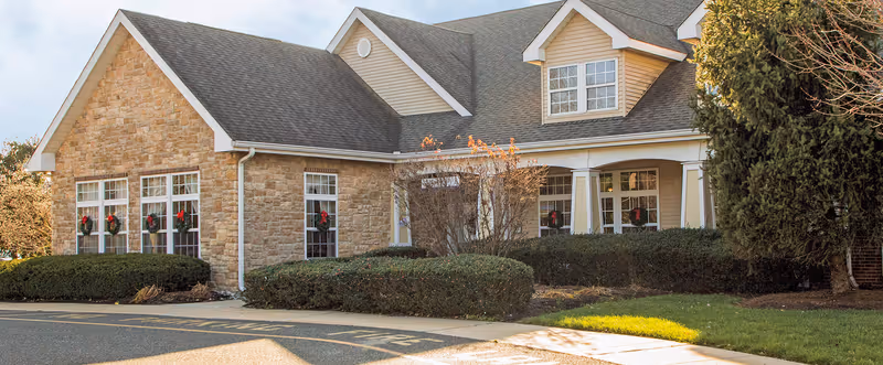 Exterior view of a senior living facility building with stone and siding walls, multiple windows decorated with holiday wreaths, a covered entrance, and well-maintained bushes and trees in the front.