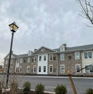 Front exterior view of a two-story brick senior living building with a lamp post and small shrubs under an overcast sky.