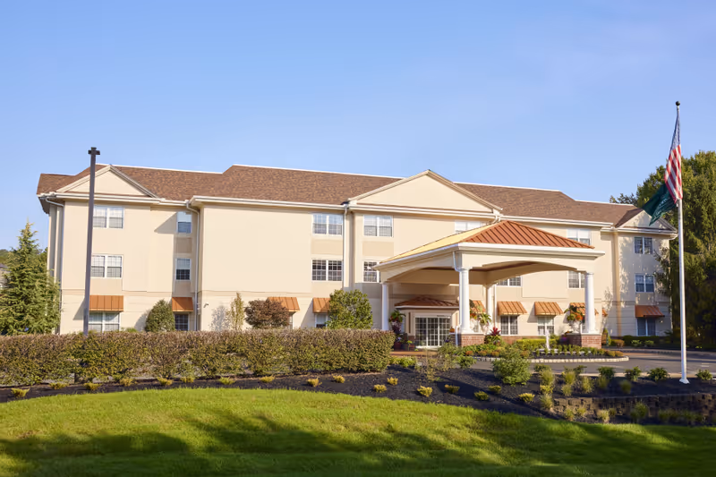 Exterior view of a three-story senior living facility building with beige walls and brown roof. The entrance has a covered portico with white columns and a brown roof. There are well-maintained shrubs, plants, and a green lawn in front of the building. An American flag and a green flag are flying on a flagpole to the right.