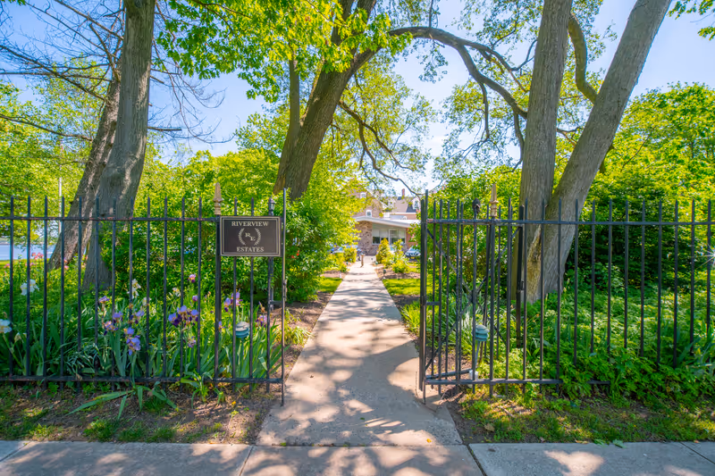 Black iron gate entrance to Riverview Estates with a concrete pathway leading through a lush garden with trees and flowers towards a building in the background under a clear blue sky.