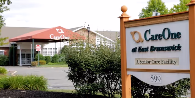 Entrance of CareOne at East Brunswick senior care facility with a red canopy over the driveway and a sign in the foreground displaying the facility name and address number 599.