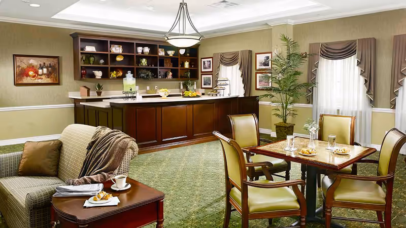 A cozy common area with a small dining table set for four with plates and glasses, a comfortable armchair with a throw blanket and a cup on a side table, and a wooden counter with shelves displaying decorative items in the background. The room has green patterned carpet, beige walls with white trim, and windows with draped curtains.
