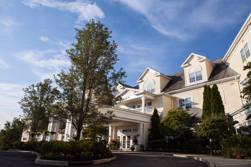 Exterior view of a large, multi-story senior living facility with beige siding, white trim, and multiple dormer windows. The entrance features a covered driveway with columns, surrounded by landscaped greenery and trees under a partly cloudy blue sky.