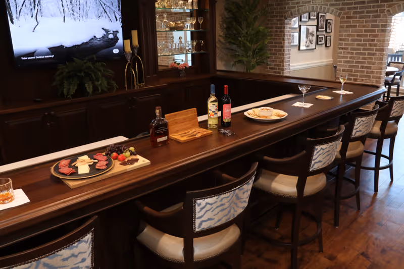 A cozy bar area with a long wooden counter and five cushioned bar stools with patterned backs. On the counter, there are plates with snacks including cheese, crackers, grapes, and strawberries, along with bottles of wine and a bottle of whiskey. Behind the bar, there is a cabinet with glassware and a television mounted on the wall showing a snowy landscape. The room features brick walls and wooden flooring, creating a warm and inviting atmosphere.