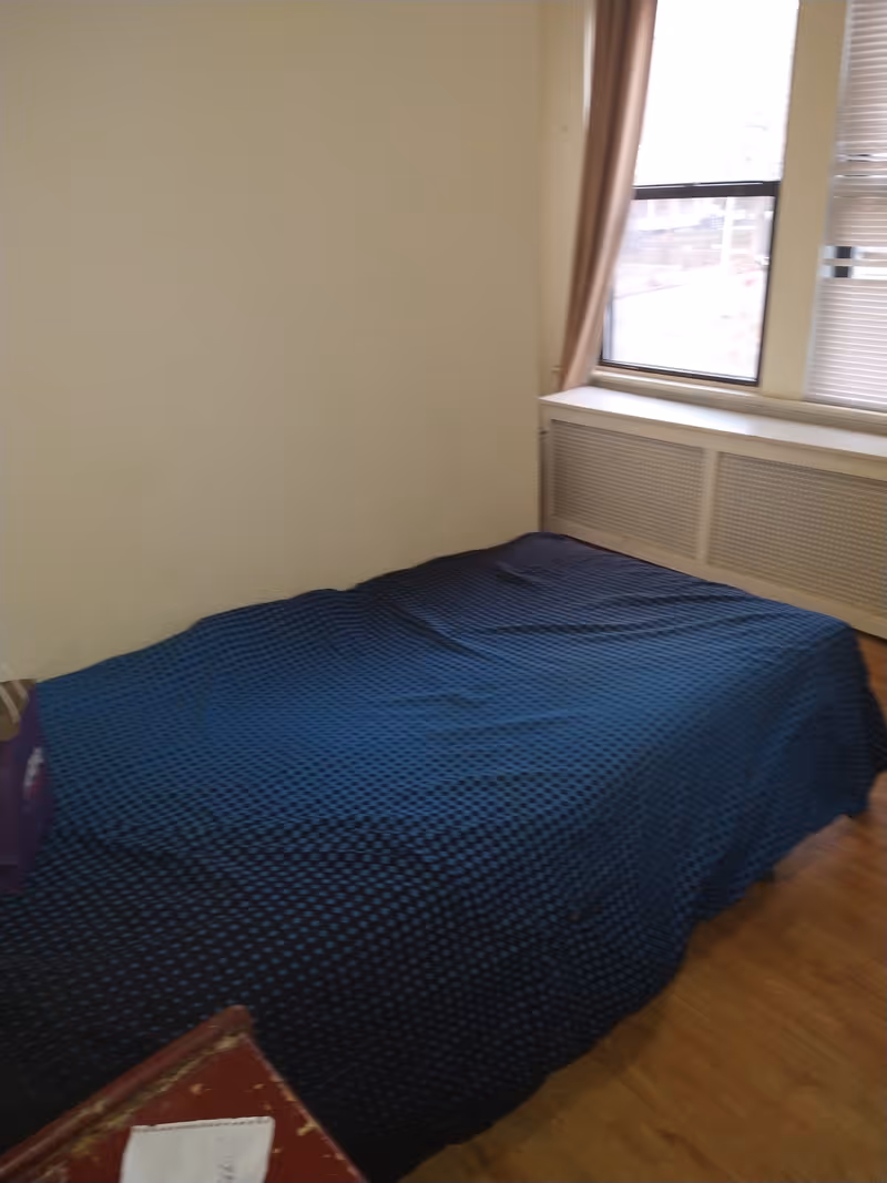 A simple bedroom with a single bed covered in a dark blue blanket with a subtle pattern. There is a window with beige curtains and blinds partially open, letting in natural light. The floor is wooden, and a small portion of a red piece of furniture is visible in the foreground.