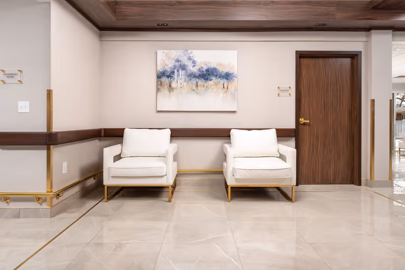 A clean and modern waiting area with two white cushioned armchairs with gold legs placed side by side against a beige wall. Above the chairs is a painting featuring abstract blue and beige trees. To the right is a wooden door with a gold handle, and small signs on the walls indicate directions to a conference room and admissions. The floor is polished beige tile with gold trim, and the ceiling has wood paneling with recessed lighting.