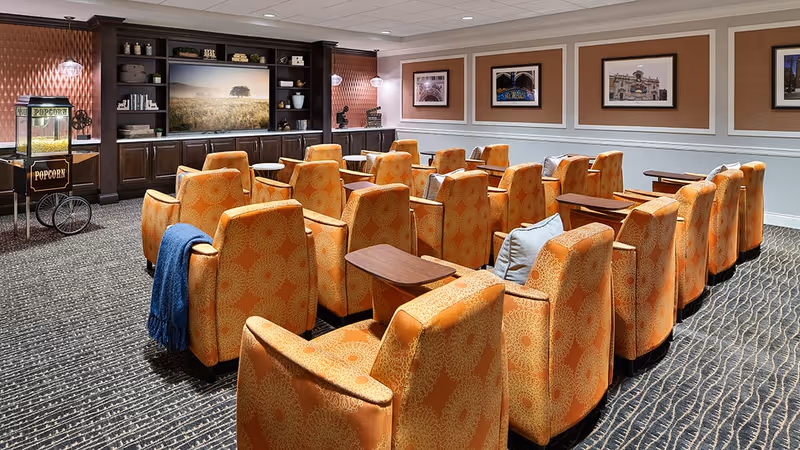 A cozy media room with multiple rows of orange patterned armchairs facing a large flat-screen TV mounted on a dark wood entertainment center. There is a popcorn machine on the left side of the room and framed pictures on the walls. The room has carpeted flooring and soft lighting.
