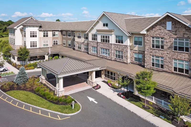 Exterior view of a large senior living facility building with multiple windows, a covered entrance with a driveway, landscaped greenery including trees and shrubs, and a clear blue sky.