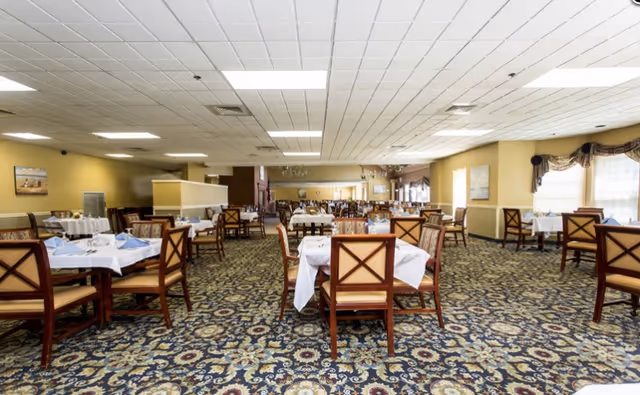 Spacious dining room with multiple tables and wooden chairs set with white tablecloths and a patterned carpet.