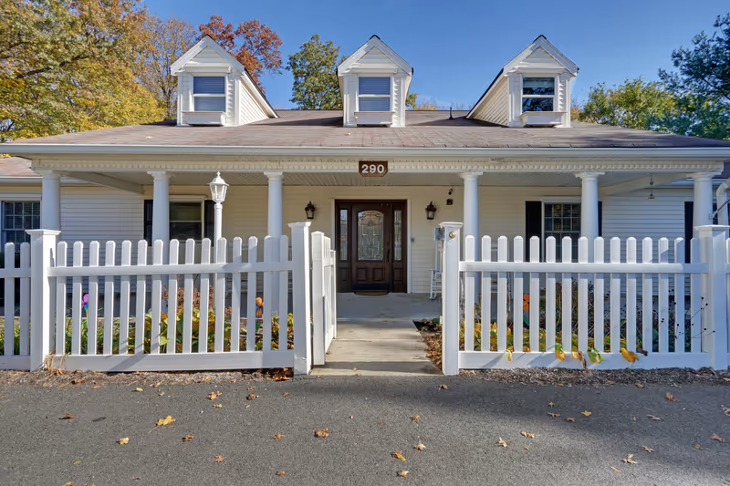 Front exterior view of a single-story building with white siding, three dormer windows on the roof, a dark wooden front door with glass panels, and a white picket fence enclosing a small garden area. The building number 290 is displayed above the door. Trees with autumn foliage are visible in the background under a clear blue sky.