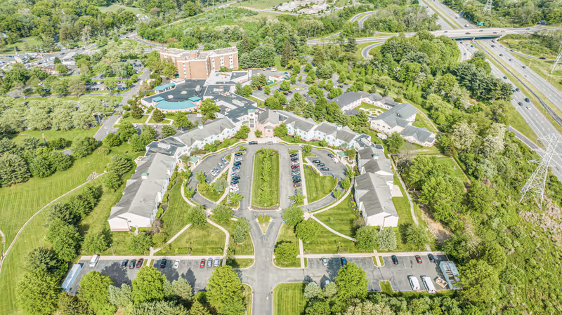 Aerial view of The Terraces at Lawrence senior living facility showing multiple buildings arranged around a central parking area with green lawns and trees. Surrounding the facility are more trees, roads, and a highway visible in the background.