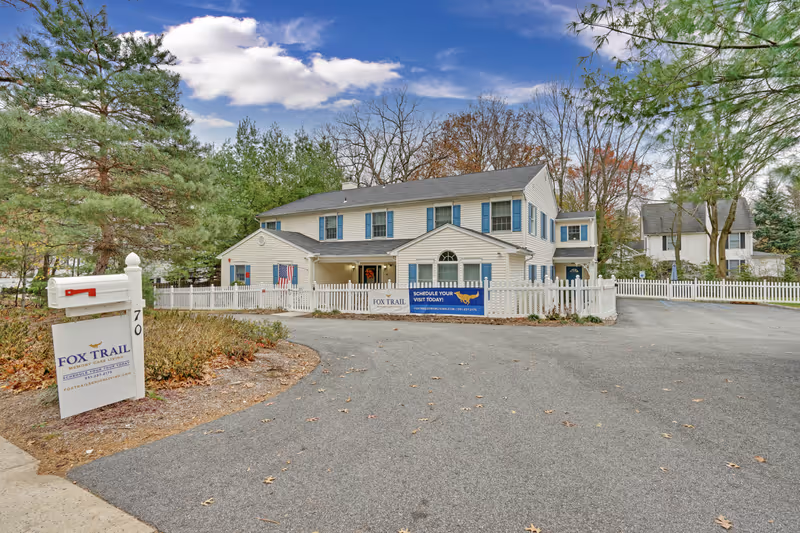 Exterior view of a two-story building with white siding and blue shutters, surrounded by a white picket fence. There is a paved driveway leading to the building, and trees with autumn foliage in the background. A white mailbox and a sign reading 'Fox Trail Memory Care Living' are visible near the driveway entrance.